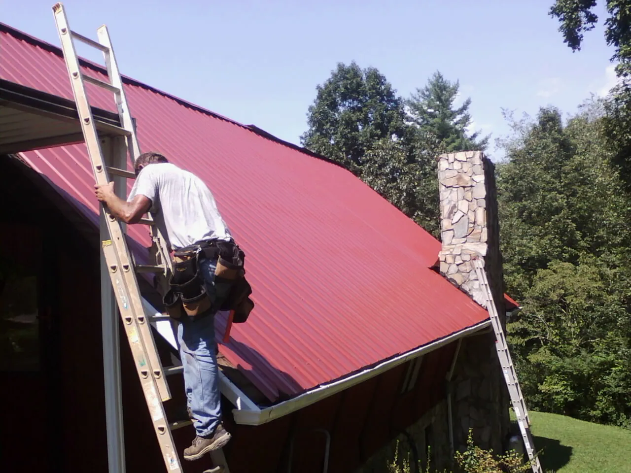 Roofer on a ladder beside a red metal roof and stone chimney — Soffit & Fascia Repair by Roofing Brothers in Dukedom